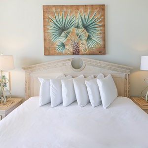 Master suite on the beach featuring a king bed adorned with white pillows, complemented by tropical art and elegant lamps