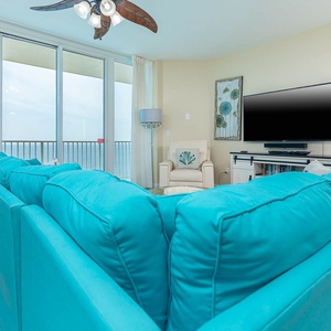 This living area features a vibrant turquoise sofa, a large TV, and stunning beachfront views through expansive windows