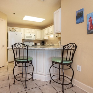 Bright kitchen area features a breakfast bar with green cushioned stools, white cabinetry, and decorative wall art