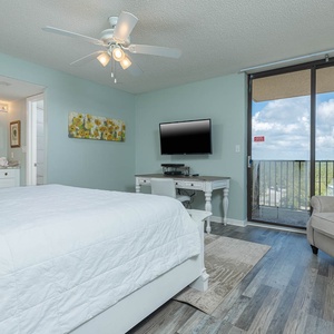 Master suite featuring a king bed, a cozy armchair, a desk with a TV, and access to a balcony with a beach view