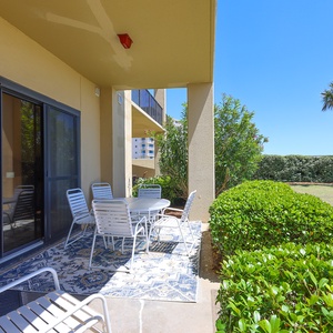 The ground floor patio features a round table with chairs, surrounded by lush greenery and palm trees under a clear blue sky