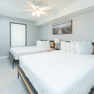 Bright guest bedroom featuring two twin beds, a bedside lamp, and coastal artwork against soft gray walls