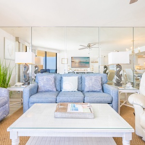Bright living area features a blue sofa with queen sleeper, cozy chairs, and panoramic views of the beach