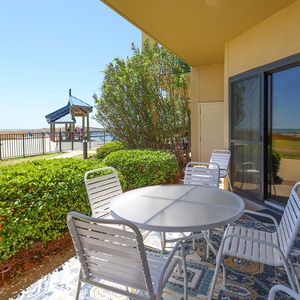 A cozy outdoor patio area features a round table and chairs, surrounded by lush greenery, the pool area, and a beach view