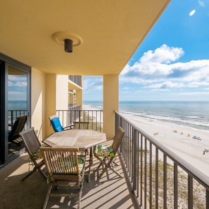 A spacious balcony features a wooden dining table and chairs, offering a panoramic view of the beach and gentle waves