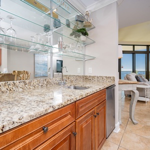 This elegant wet bar features a granite countertop, a blender, glassware, and a view of the inviting living area and beach