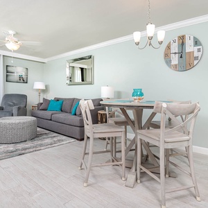 The living area features a gray sofa with queen sleeper, a round coffee table, and a dining set with a beach view