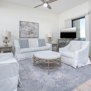 Bright living area features white sofas, a round coffee table, and a TV, with large windows offering a glimpse of the gulf