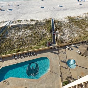 The view captures the sparkling pool area with lounge chairs, leading to a serene stretch of white sand beach