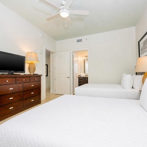 This guest bedroom features two comfortable beds, a wall-mounted TV, and a cozy lamp beside a stylish dresser