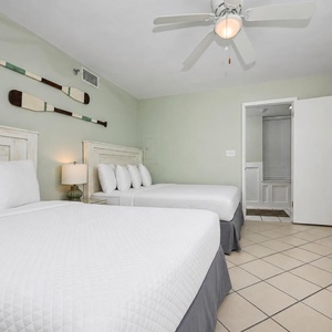 This guest bedroom features two queen beds with white linens, a ceiling fan, and decorative oars on the green wall