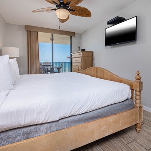 The inviting master bedroom features a wooden king bed, a ceiling fan, and a balcony view of the beach, complemented by a TV