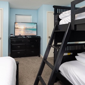 A cozy guest room features a black full over full bunk bed, a queen bed, a dresser, and a TV displaying a serene beach scene