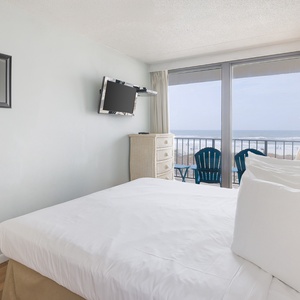 The bedroom features a cozy bed, a TV, and a stunning beachfront view from the balcony seating area