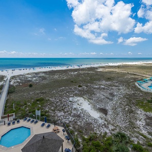 Expansive beach front view reveals white sand and vibrant blue waters, with a pool area and lounge chairs inviting relaxation