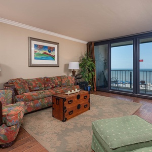 The living area features colorful floral sofas, a wooden coffee table, and a stunning beach view through large windows
