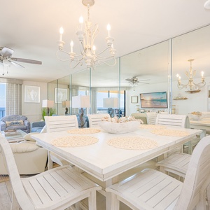 Bright dining area with white wooden furniture, a chandelier, and views of the beach through large windows