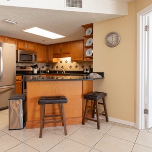 Modern kitchen features wooden cabinetry, a black countertop, and a breakfast bar with two stools, creating a welcoming space