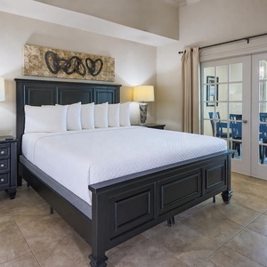 The master bedroom features a plush king bed with crisp white linens, elegant decor, and a view through into the dining area