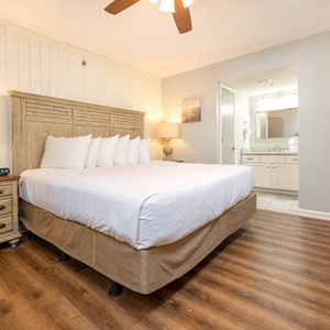 Brightly lit master suite featuring a king bed with plush pillows, a wooden nightstand with a lamp, and an adjoining bathroom