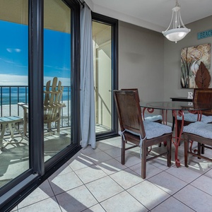 The dining area features a glass table with wooden chairs, offering a view of the beach through large sliding doors