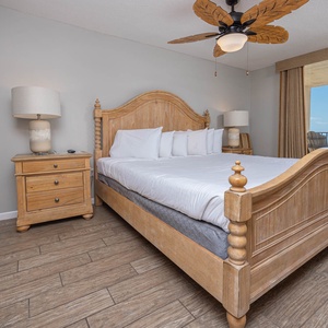 The cozy master bedroom features a wooden king bed, plush bedding, and a balcony view with seating overlooking the beach