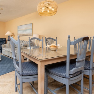 Dining area features a glass-top table with blue cushioned chairs, complemented by a cozy living space and beach view beyond