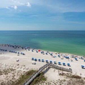 A vibrant beach scene features colorful umbrellas, sunbathers, and a wooden walkway leading to the pristine shoreline