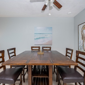 The dining area features a rustic wooden table surrounded by dark leather chairs, complemented by beach-themed artwork