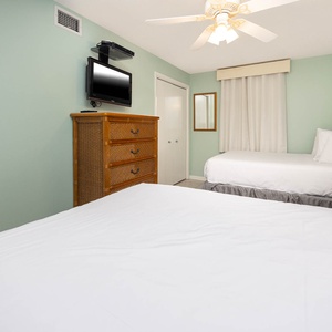 A serene guest bedroom features two double beds, a wooden dresser, a mounted TV, and green walls for a calming atmosphere