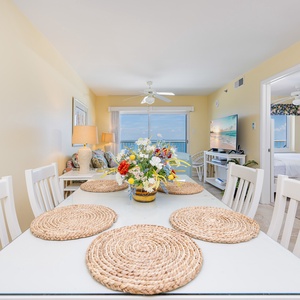 The dining area features a bright table set with woven placemats, a lovely floral centerpiece, and a view of the beach