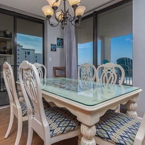 Bright dining area features a glass-top table with elegant white chairs and a view of the beach through large sliding doors