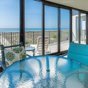 Bright seating area with a glass table offers a serene view of the beach and gulf from this enclosed balcony area
