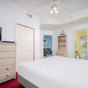 Bright bedroom featuring a plush king bed, dresser with TV, and a glimpse of an inviting living area through the doorway