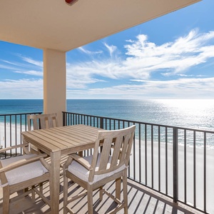 The balcony features a wooden dining set with cushioned chairs, offering a panoramic view of the beach and sparkling water