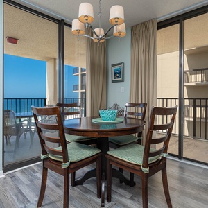A round dining table with wooden chairs and a decorative bowl is set against large windows offering a beach view