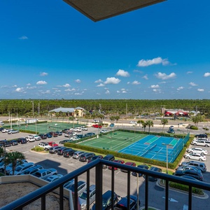 The view from the balcony showcases vibrant tennis courts and a bustling parking area under a clear blue sky