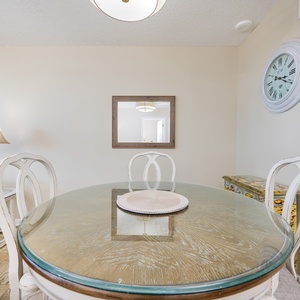 A round glass dining table with white chairs is complemented by a lamp and a decorative clock against a neutral wall