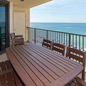 The balcony features a wooden dining table with chairs, offering a direct view of the vibrant beach and turquoise waters