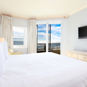 This bedroom features a plush king-size bed, a wall-mounted TV, and stunning beachfront views through large sliding doors