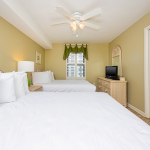Bright guest bedroom featuring two queen beds, a TV, and a window with green accents, creating a cheerful atmosphere