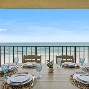 A dining table set for six features elegant blue glassware and white plates, with a serene beachfront view in the background