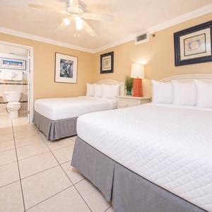 Bright guest bedroom featuring two double beds, a colorful lamp, and artwork, with a doorway leading to a bathroom area