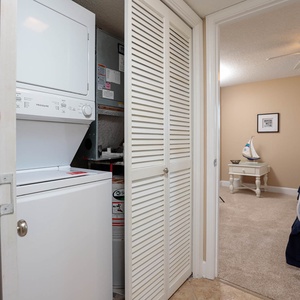 A stacked washer and dryer are visible in a closet, leading to a cozy guest bedroom with two beds and light beige walls