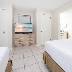 Bright guest room featuring two double beds, a TV on a wooden dresser, and a doorway leading to a well-lit bathroom area