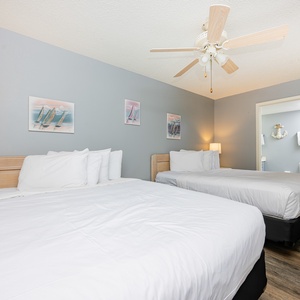 This guest bedroom features two queen beds, coastal artwork, a ceiling fan, and a warm ambiance with natural wood accents