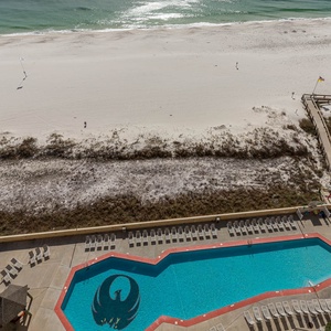 The view showcases a sparkling pool area with lounge chairs, leading to a pristine beach with soft white sand