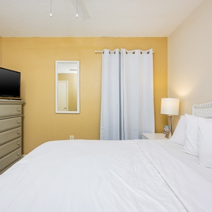 This guest bedroom features a comfortable queen bed, a TV, and cheerful beach-themed artwork on warm yellow walls