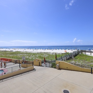 The balcony view showcases the vibrant pool area with red lounge chairs, leading to a pristine beach and clear blue skies