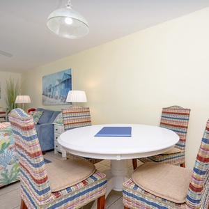 A round dining table with colorful woven chairs complements the cozy living area, featuring beach-themed artwork and lamps